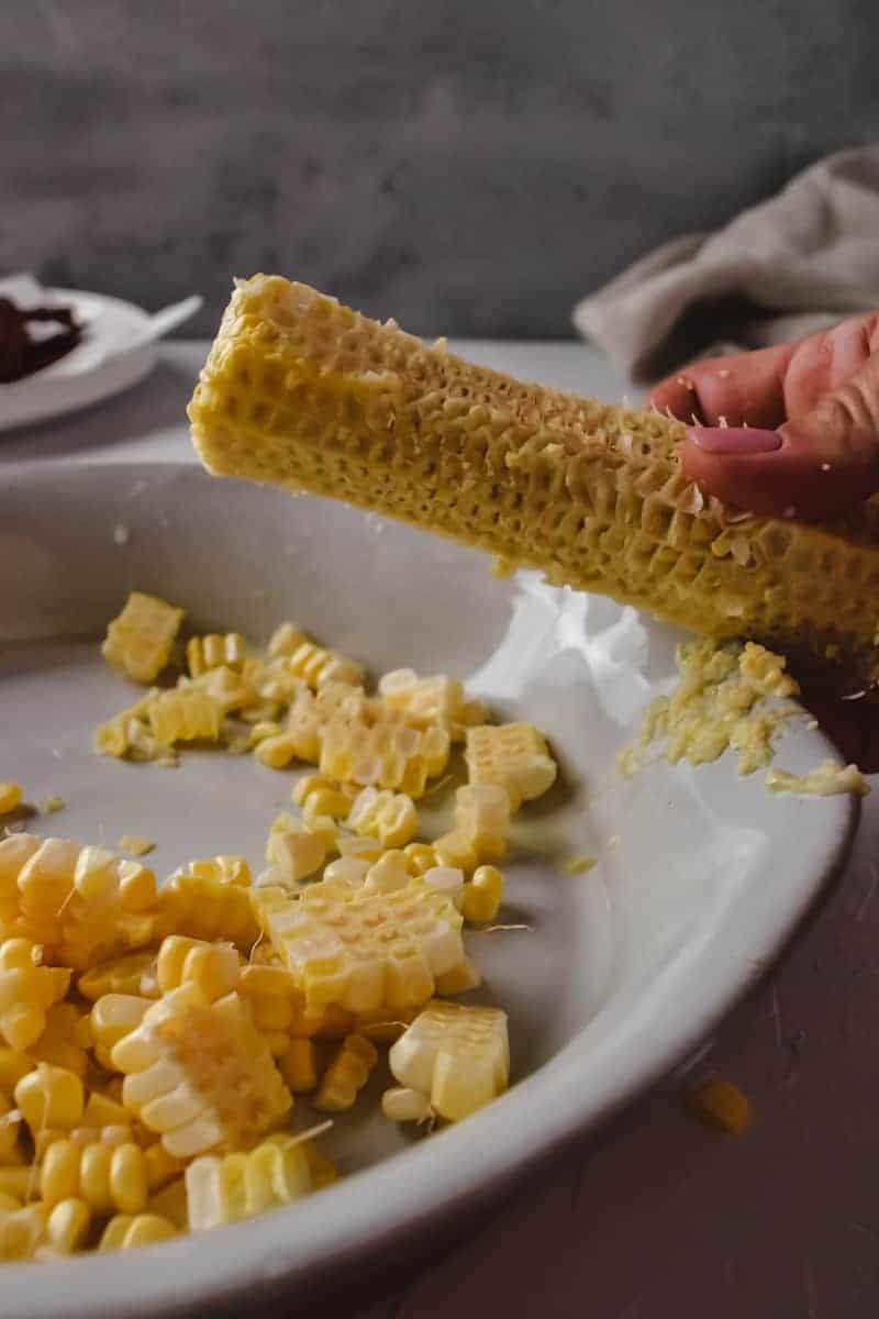 An ear of corn with kernels removed being scraped on the side of a baking dish to remove corn milk. 