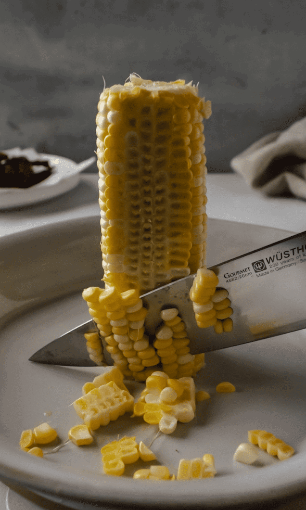 An ear of corn standing upright inside a baking dish with a knife cutting kernels of the cob. 