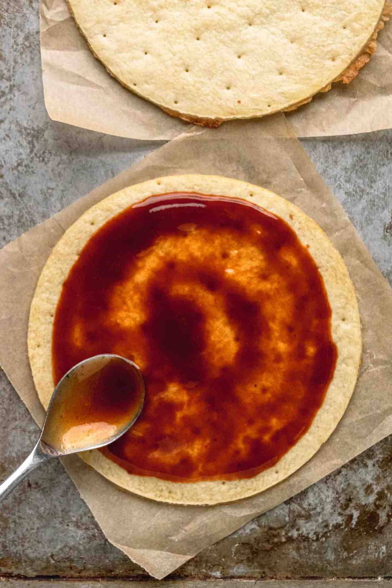 Pizza crust on baking sheet on with BBQ sauce being spread with the back of a spoon.