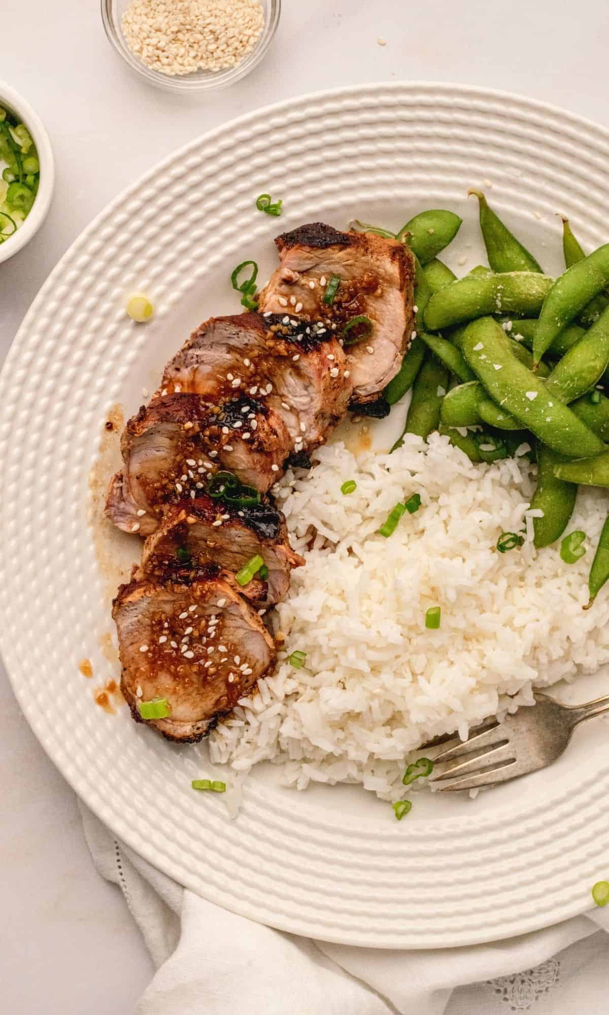 Pork tenderloin with a honey garlic sauce on a white plate with rice and edamame on the side.