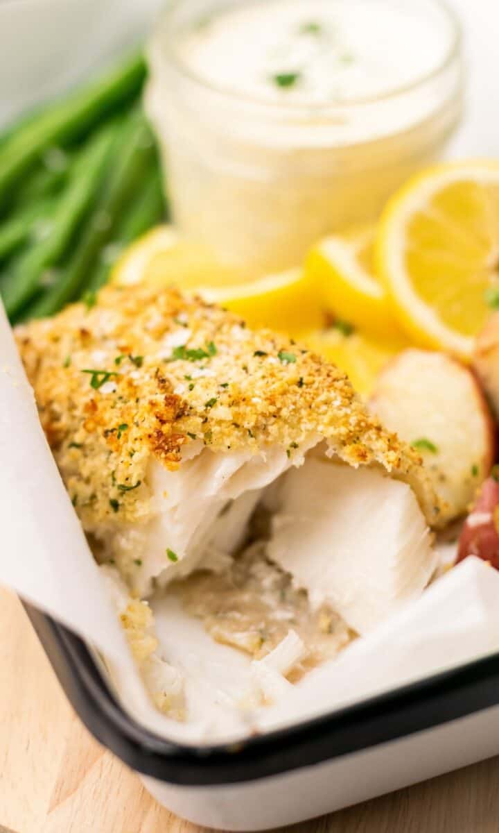 Crunchy Baked Cod with Parmesan Panko Breadcrumbs on white parchment paper surrounded by potatoes, lemon wedges and green beans.