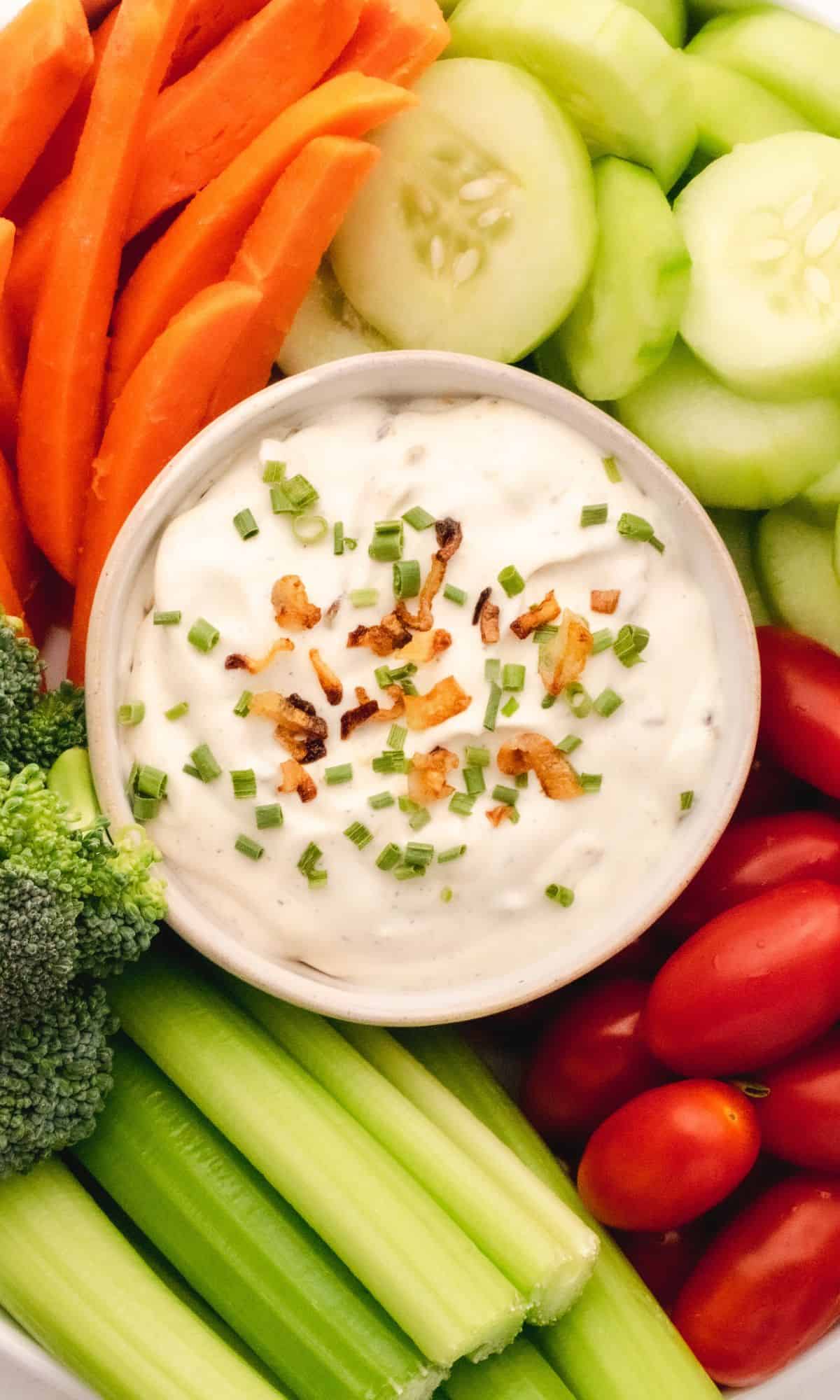 Creamy veggie dip in a white bowl surrounded by fresh veggies and topped with chives and crispy shallots.