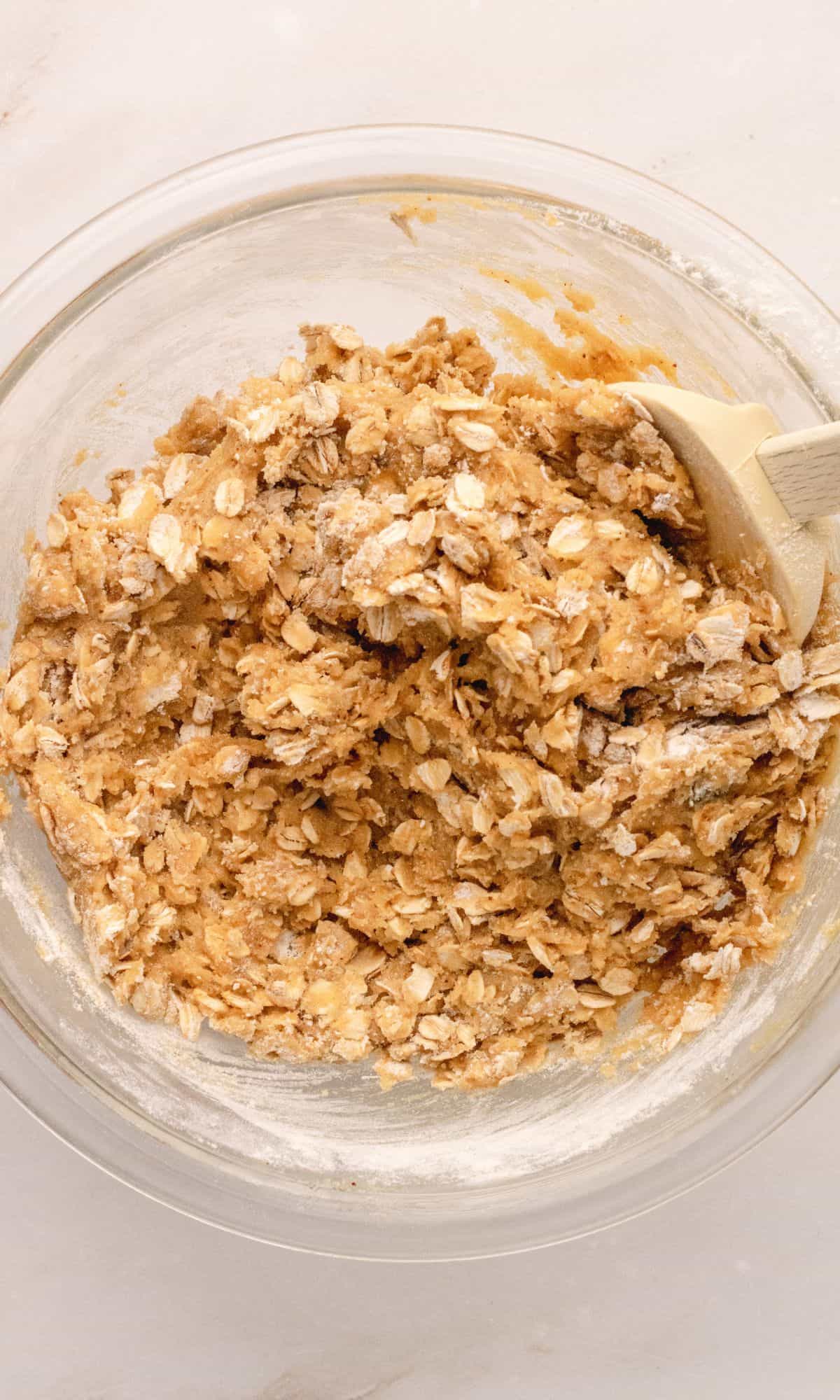 Oatmeal cookie preparation in a glass bowl being stirred with a white spatula.