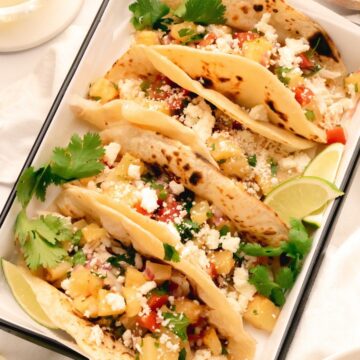 Shredded chicken tacos with roasted pineapple salsa topped with queso fresco and surrounded by cilantro and lime wedges.