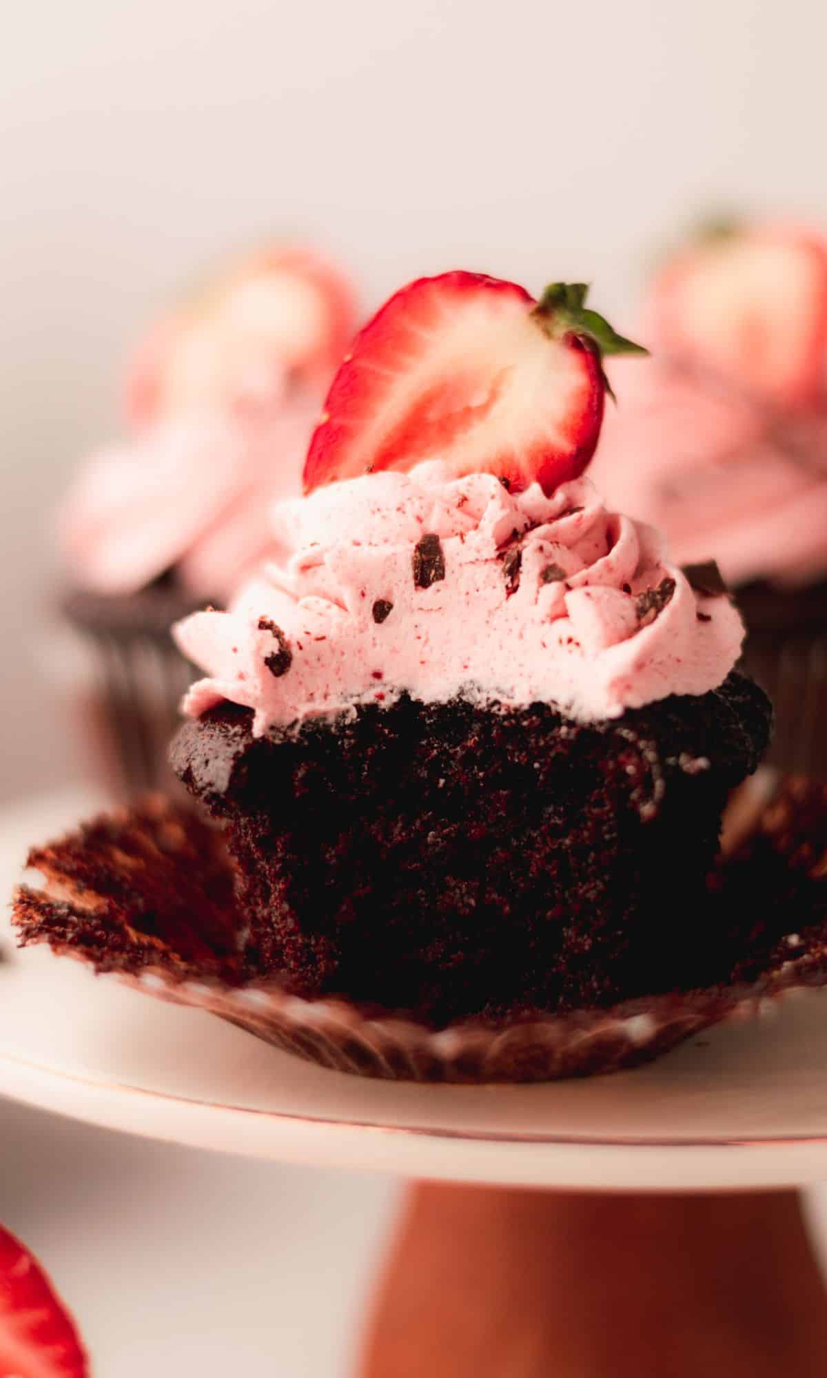 Chocolate cupcake with strawberry buttercream and a halved strawberry on top and a bite taken.