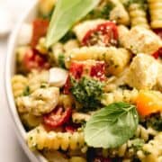 Pesto pasta salad with tomatoes and basil leaves in a white bowl.