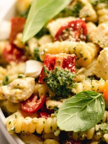 Pesto pasta salad with tomatoes and basil leaves in a white bowl.
