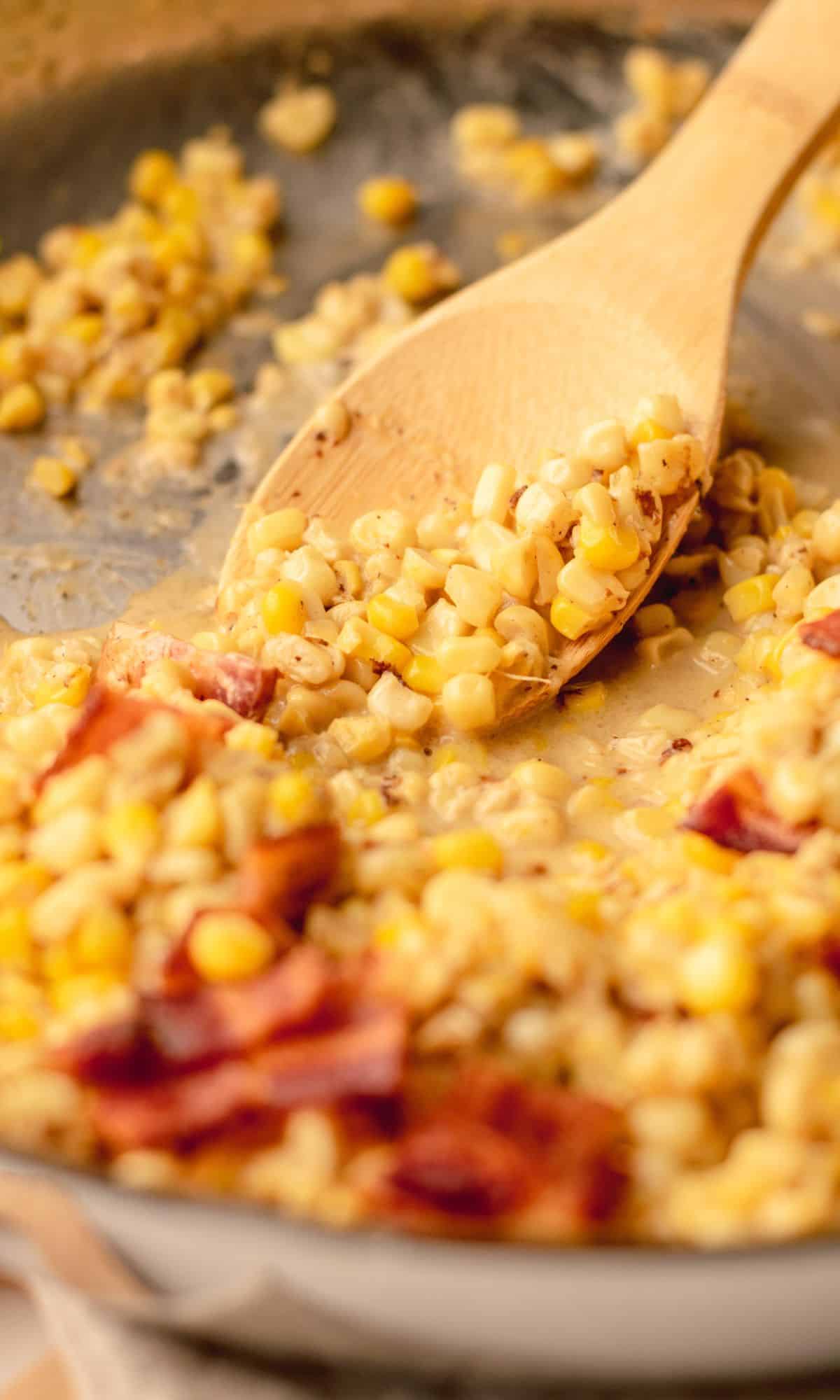 Creamy skillet corn in a skillet topped with bacon and wooden spoon scooping up corn.