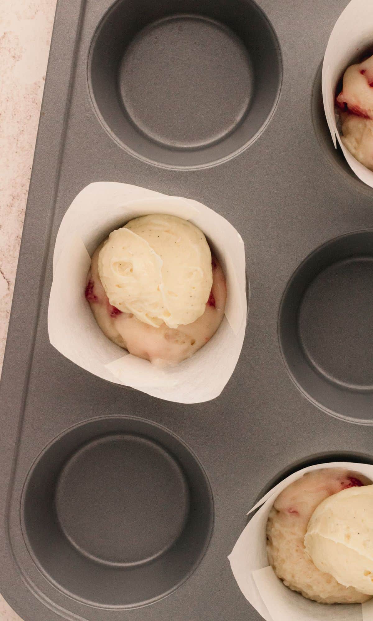A muffin tin with muffin liners and strawberry cheesecake muffin batter and cheesecake filling in the bottom.