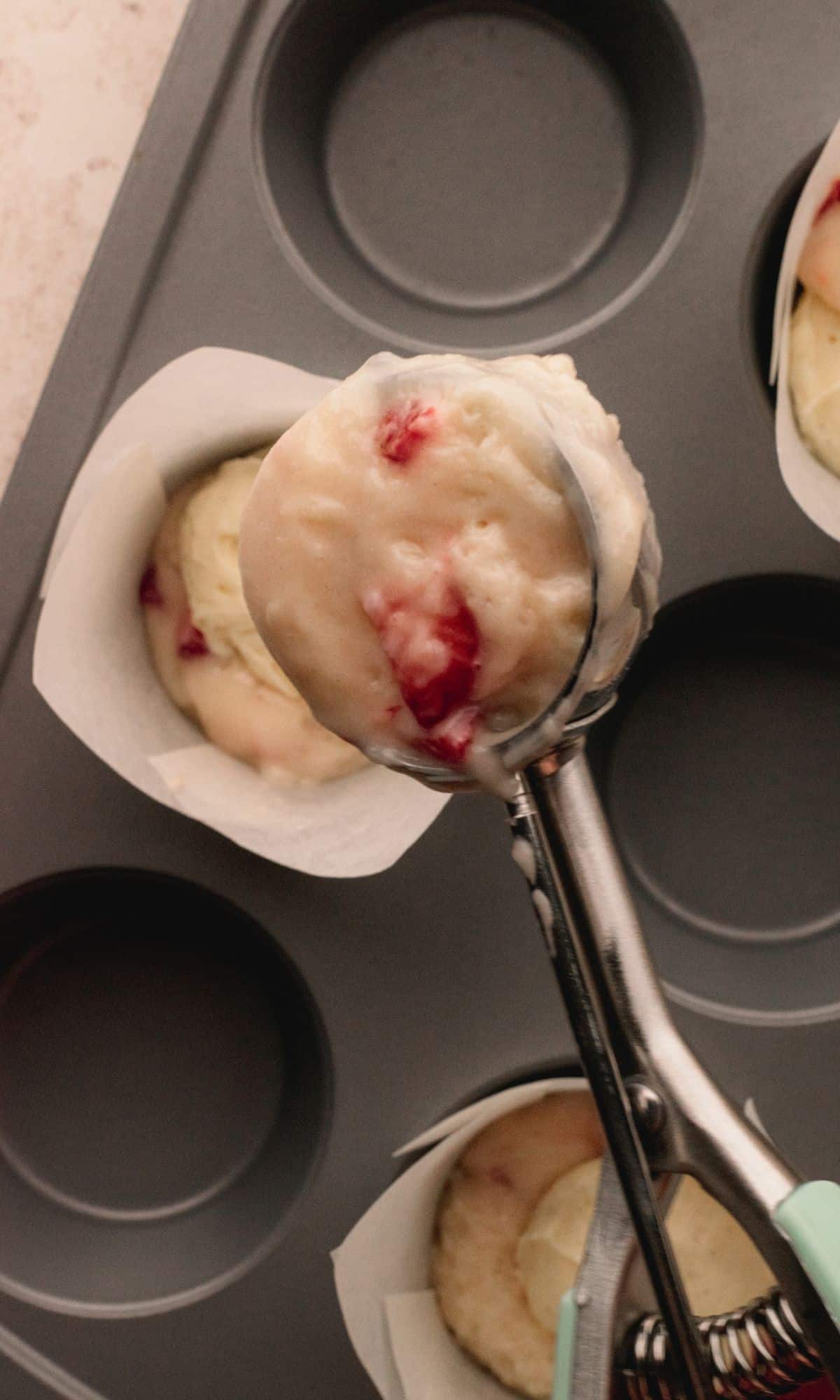 A muffin tin with muffin liners and strawberry cheesecake muffin batter in the bottom and a scoop of batter going on top.