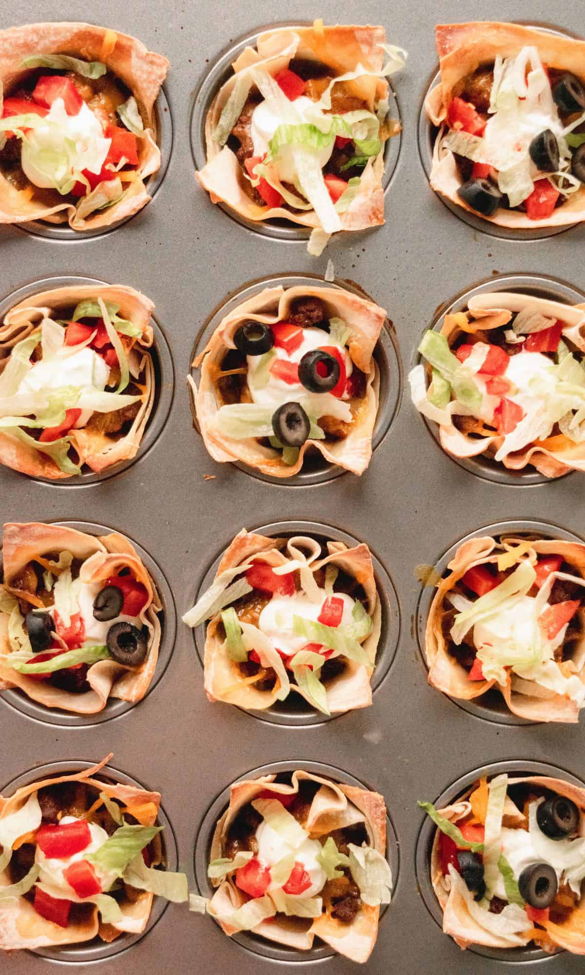 Double layer beef taco cups in a muffin tin topped with sour cream, lettuce, tomatoes, and black olives.