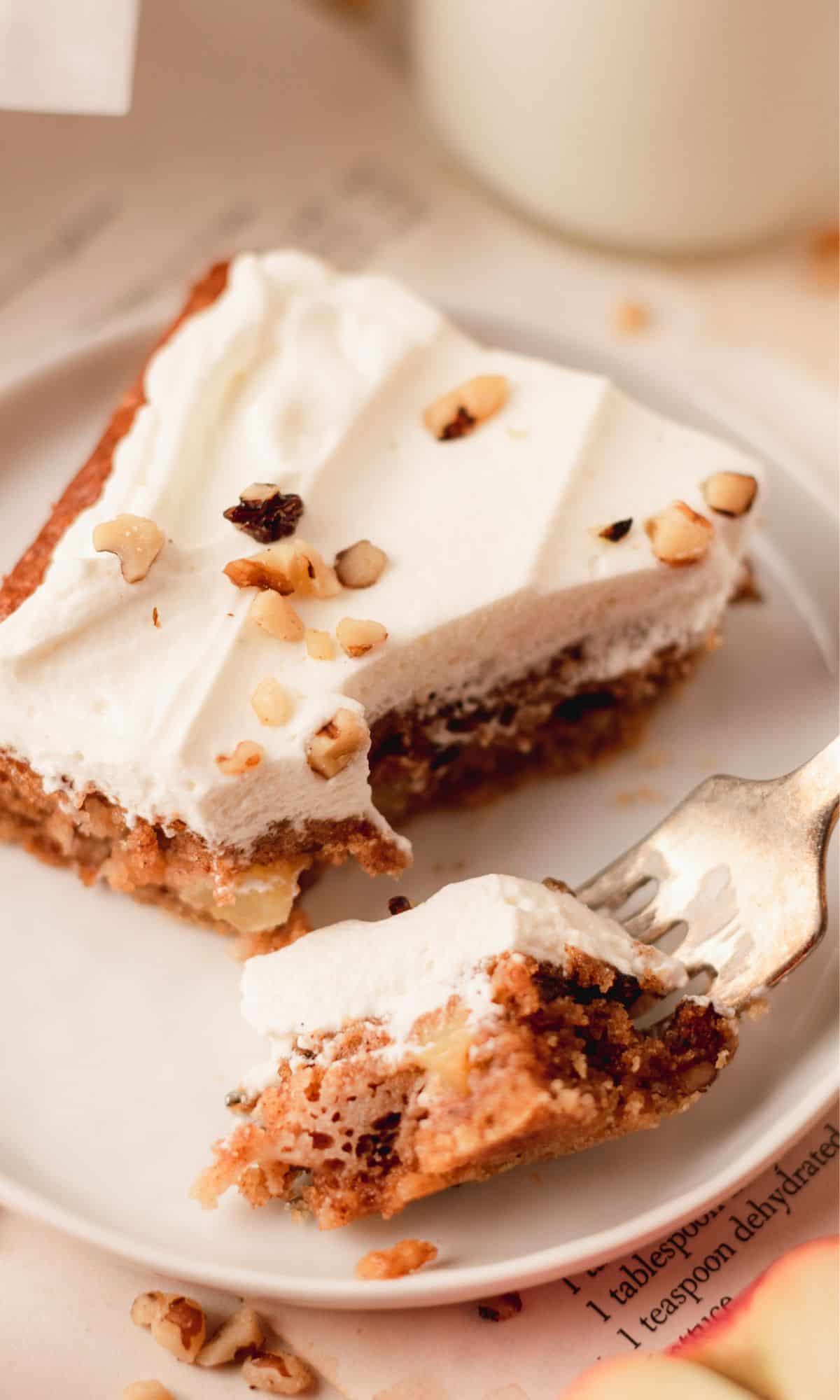 Sour cream apple bar topped with whipped cream and walnuts on a white plate with a bite on a fork.