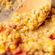 Creamy skillet corn in a skillet topped with bacon and a wooden spoon scooping up some corn..