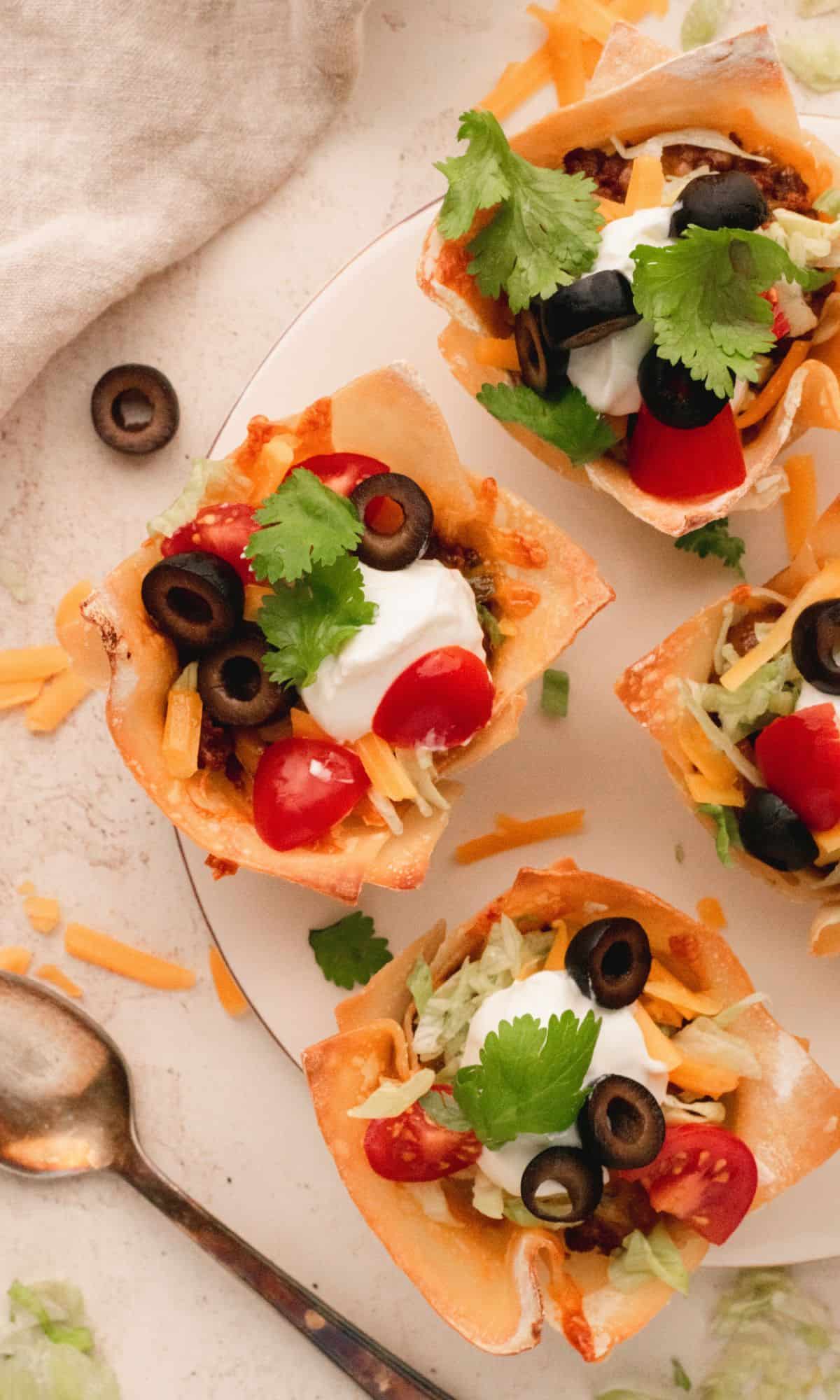 Double Layer Beef Taco Cups on a white plate topped with tomatoes, black olives, sour cream, and cilantro.