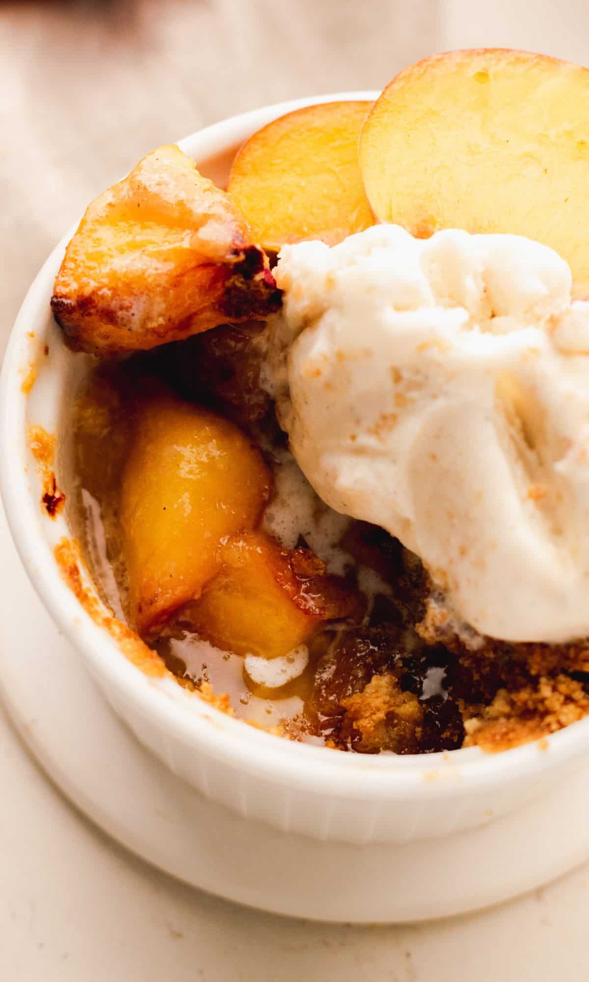 Air fryer peach crisp in a white ramekin topped with ice cream and peach slices.