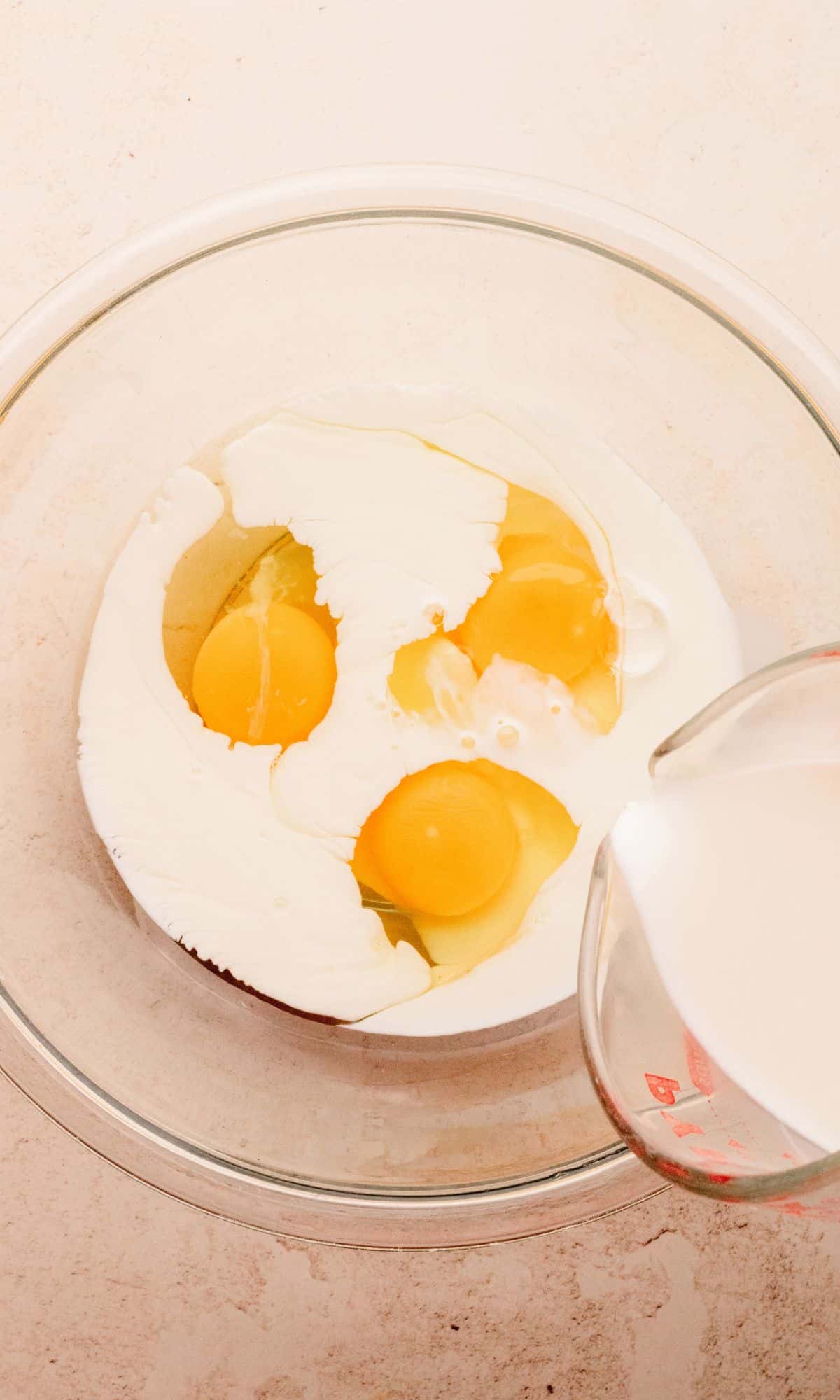A glass bowl with eggs and half and half being poured in.