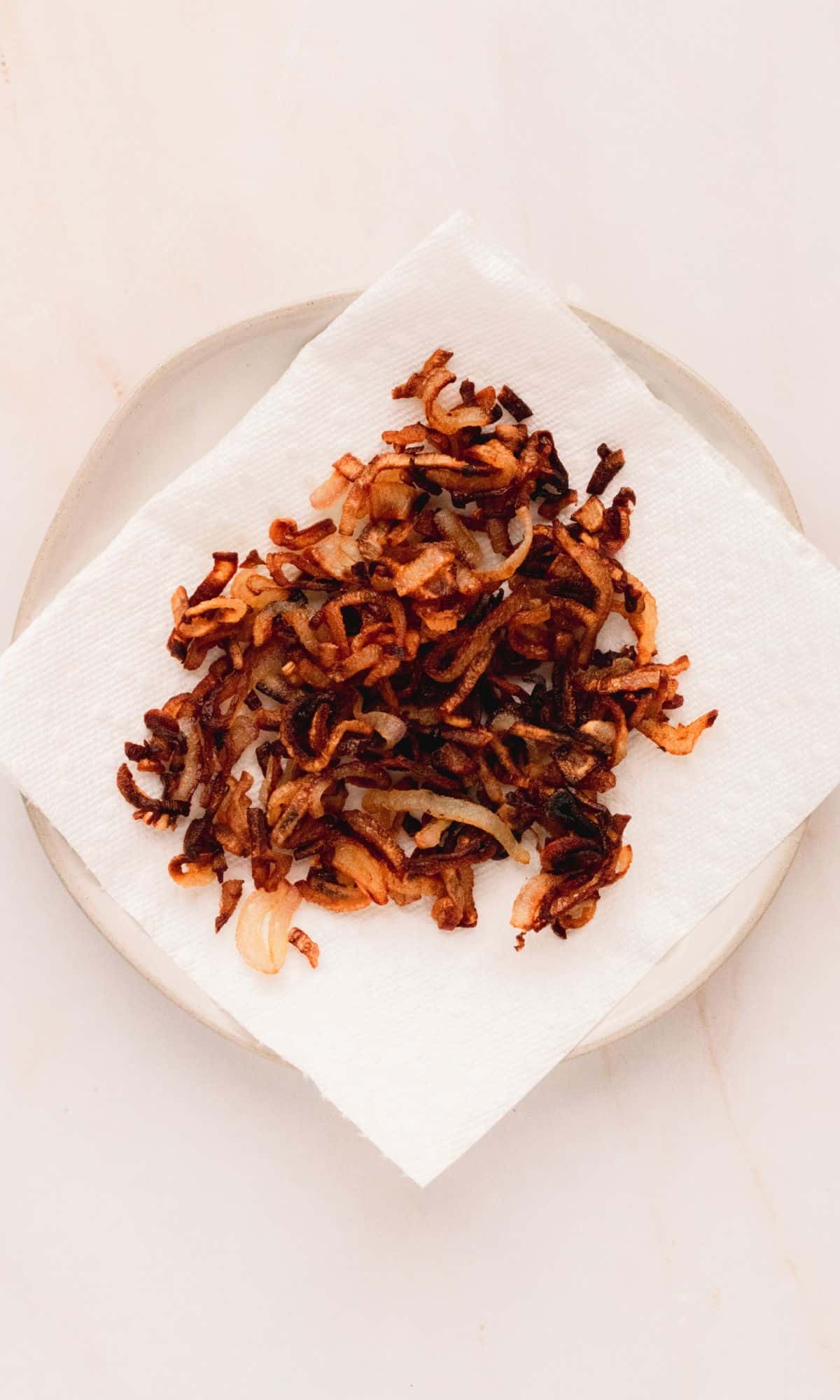 Crispy shallots draining on a white paper towel on a white plate.