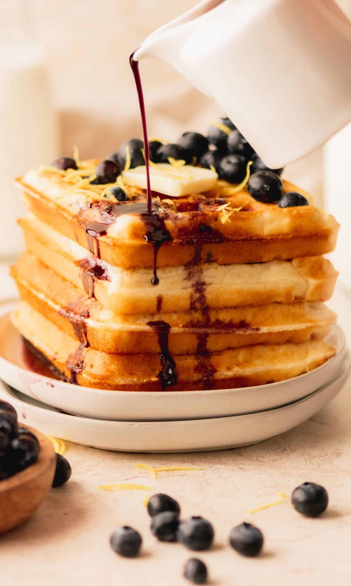 A stack of lemon waffles getting a drizzle of blueberry syrup.