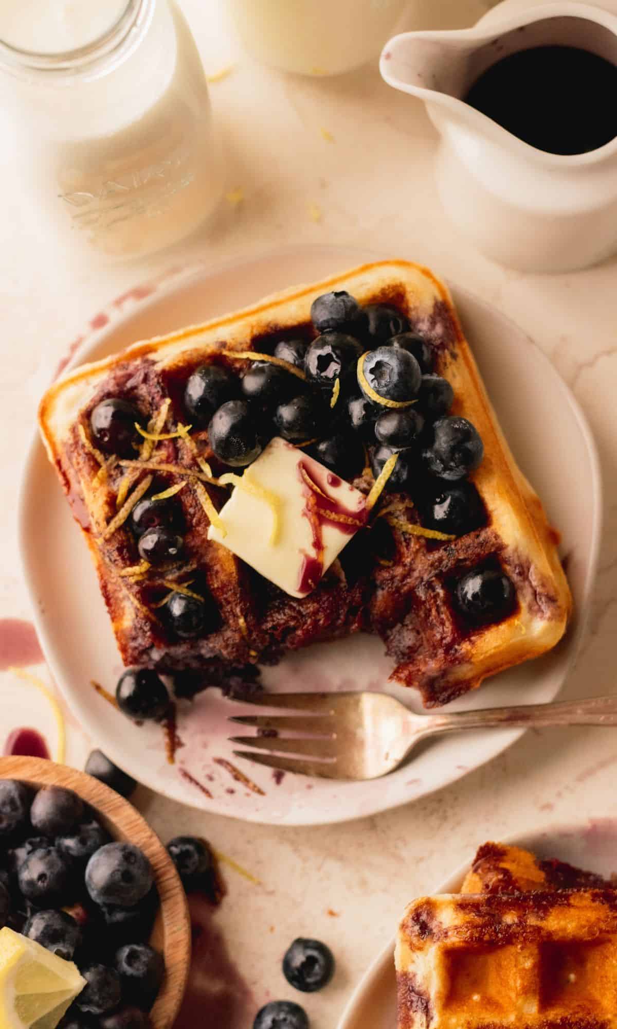 An overhead shot of lemon waffles on a white plate topped with blueberries, syrup and butter.