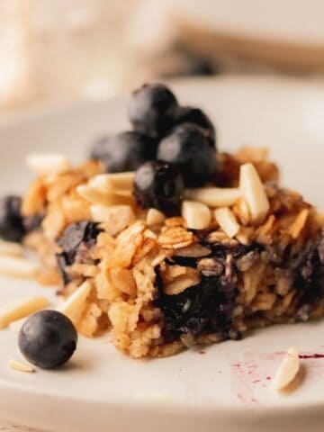 A slice of baked oatmeal on a white plate topped with blueberries and almonds.