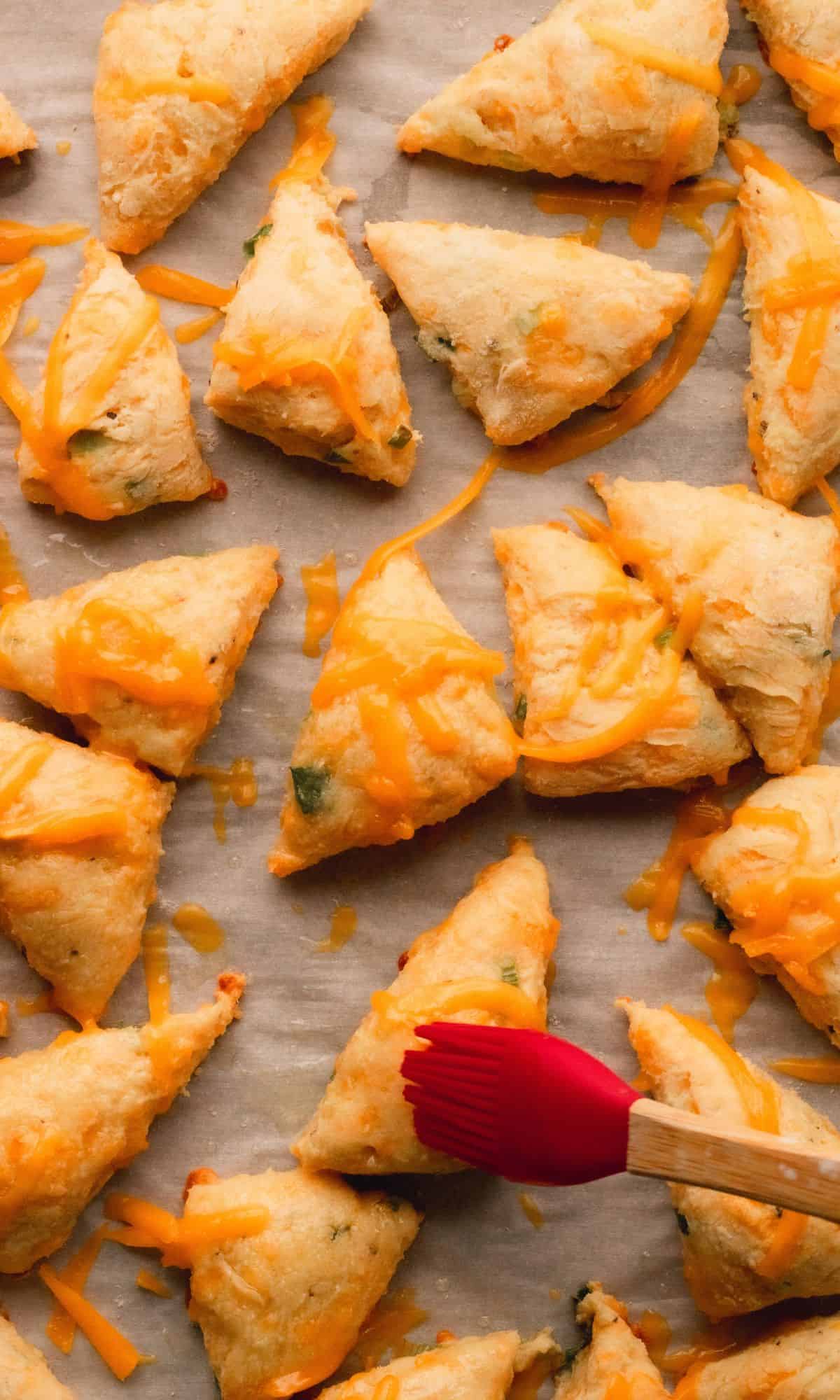 Mini cheddar scones on a baking sheet getting brushed with garlic butter.