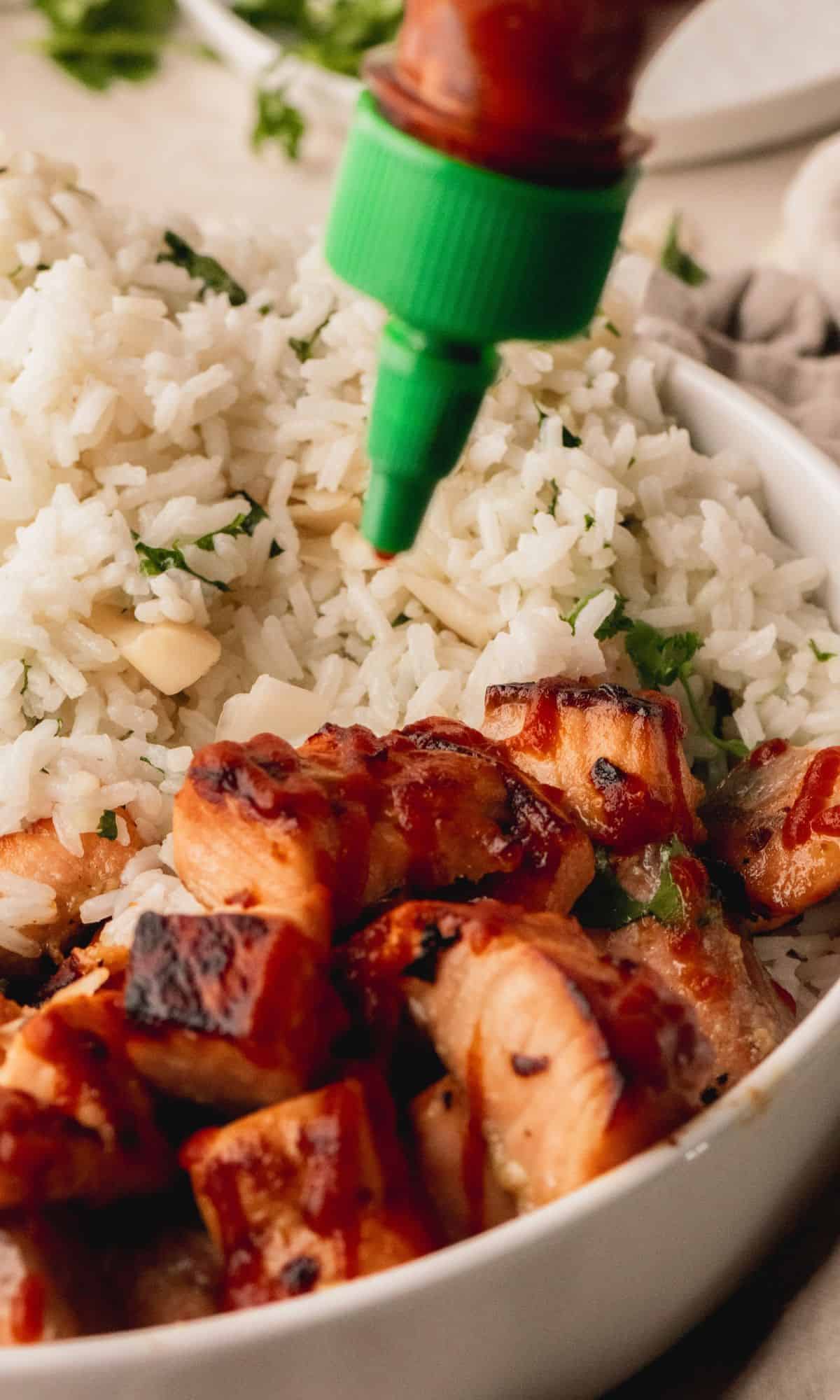 A white bowl with salmon bites, rice and broccoli getting a drizzle of Sriracha sauce.