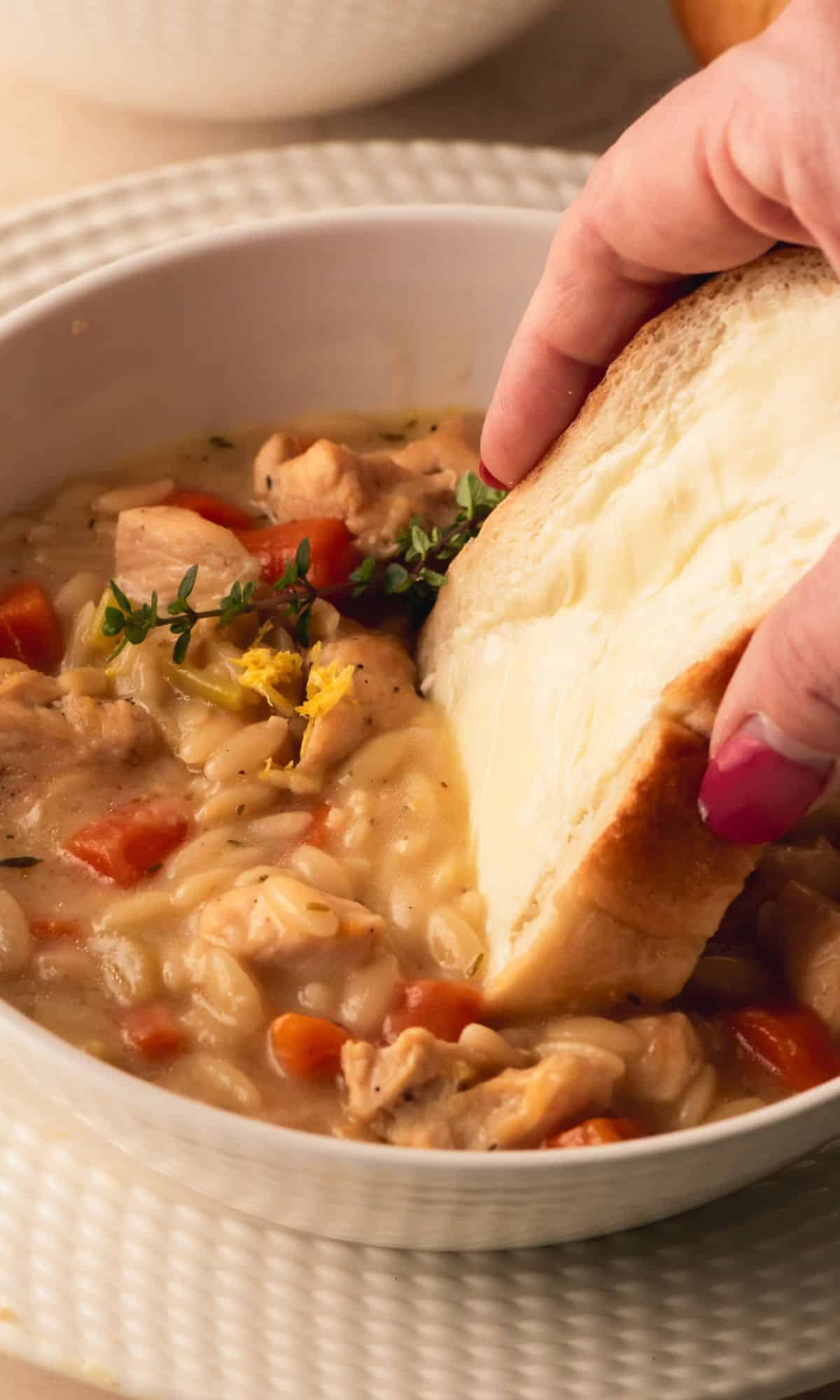 Chicken orzo soup in a white bowl with buttered bread getting dipped in.