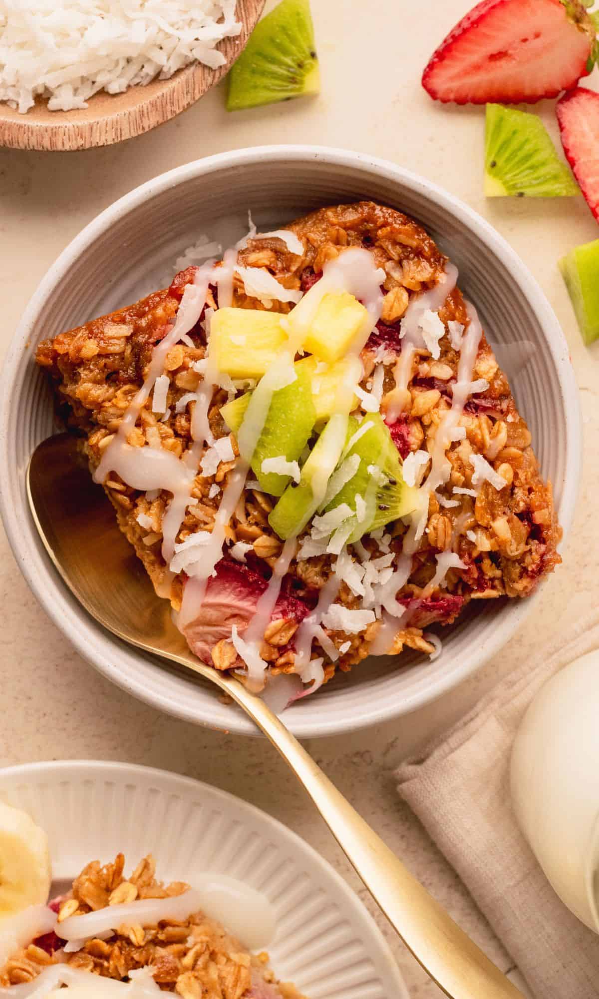 A slice of baked oatmeal in a white bowl and topped with fruit and coconut.