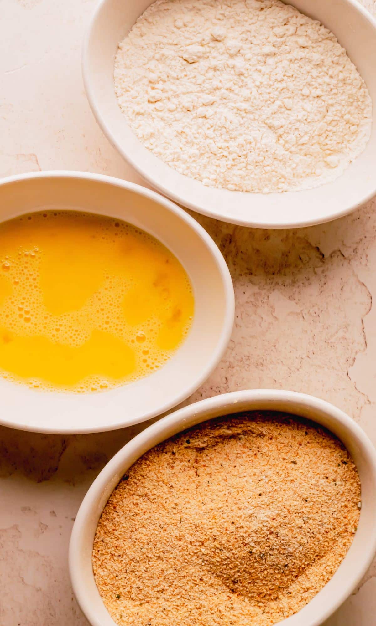 Three bowls with dredging ingredients for pork chop sandwich; one with breadcrumbs, one with egg and one with flour.