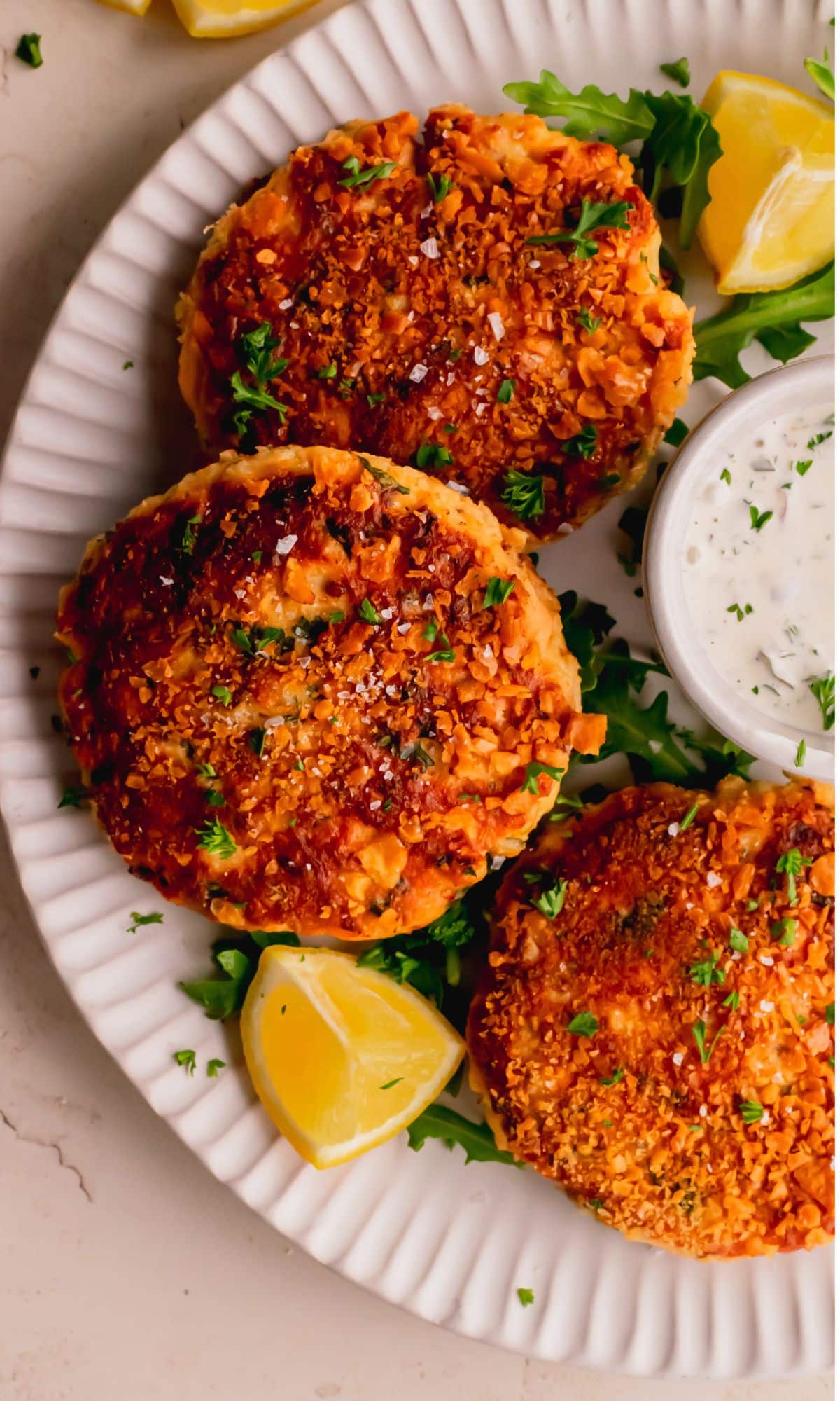 Three salmon cakes on a white plate surrounded by lemon wedges and a bowl of tartar sauce.