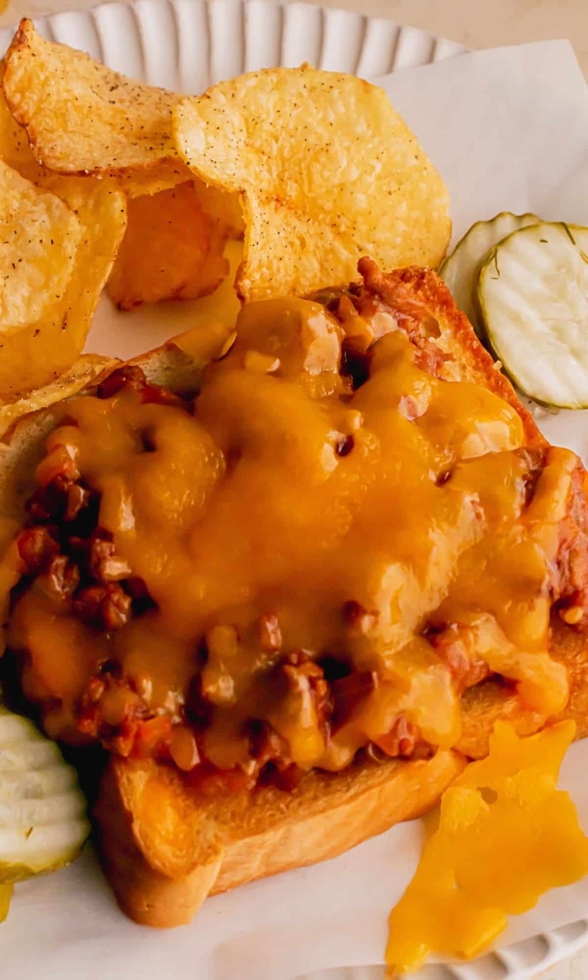 An open-faced sloppy joe on a plate with potato chips and pickles.